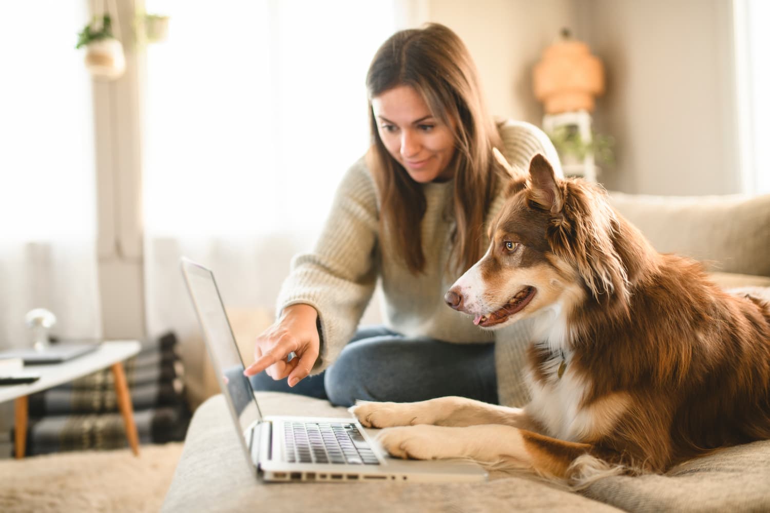 A woman sits on a couch next to a dog with its paws on a laptop as she points at the screen.