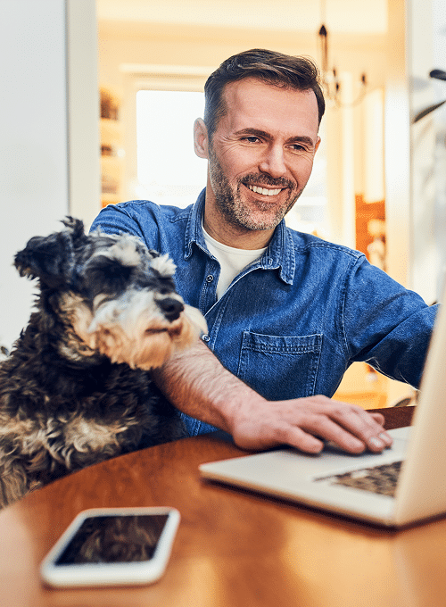 Man sitting at a table using a laptop with a dog next to him, and a smartphone lying on the table in the foreground.