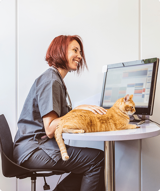 A woman in scrubs sits at a desk with a computer, smiling and petting an orange cat lying on the desk.